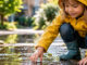 Kind in gelber Regenjacke untersucht eine Wasserpfütze im Grünen. KI-generiertes Bild.