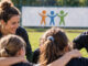 Jugendliche stehen gemeinsam mit einer Trainerin und einem Trainer im Kreis auf einem Sportplatz. Die Gruppe vermittelt Gemeinschaft, Vertrauen und gegenseitige Verantwortung. KI-generiertes Bild.