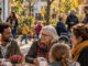 Menschen unterschiedlichen Alters und vielfältiger Hintergründe begegnen sich an einem Tisch in einem herbstlichen Sozialraum. Im Hintergrund sind weitere Erwachsene, Kinder und eine Person im Rollstuhl zu sehen. KI-generiertes Bild.