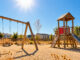 Menschenleerer Spielplatz ohne Sonnenschutz in der glühenden Mittagssonne mit trockener, spärlicher Vegetation. KI-generiertes Bild.